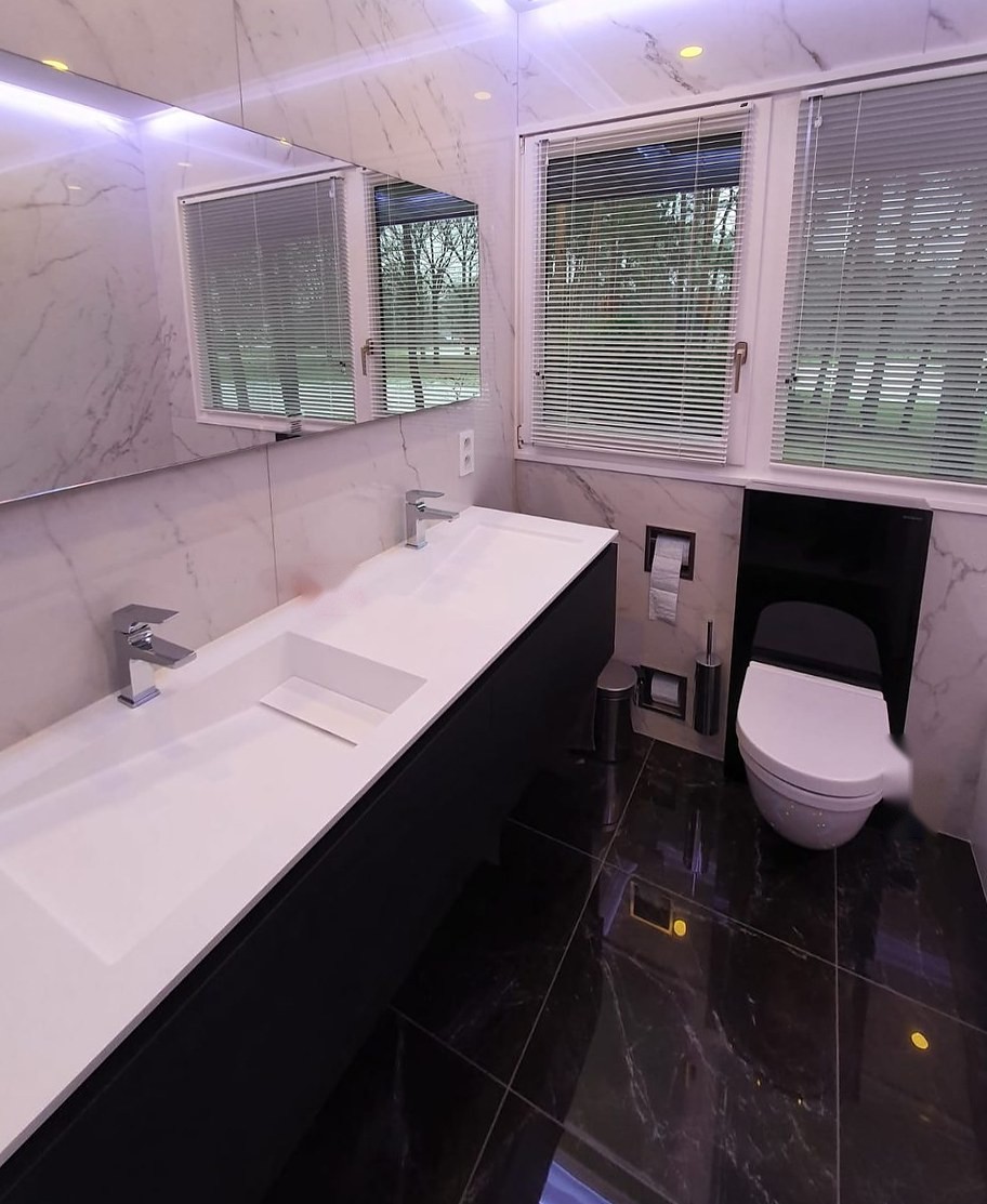 Lange witte wastafel, hangtoilet en zwarte marmerlook vloer in een smalle badkamer