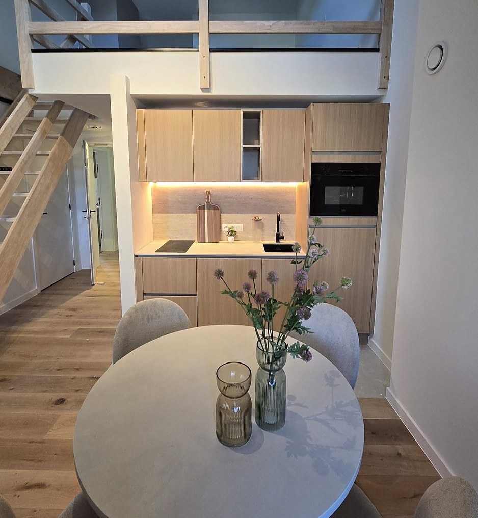 Compacte keuken in licht hout onder een mezzanine, met ronde eettafel
